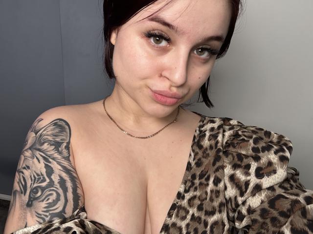 LucieRed with puffy lips, green eyes, and a tiger tattoo on her right boob, wearing a leopard print top, and a thin chain necklace.