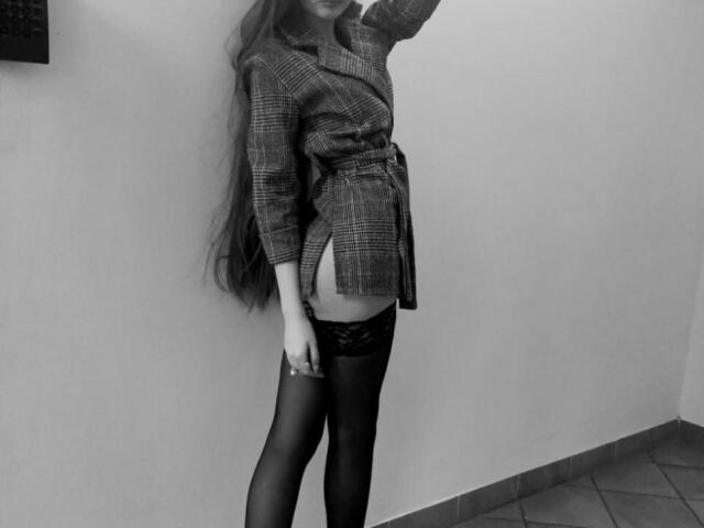 ElzaKomx in a plaid shirt, black lace stockings, and no pants, exposing her ass, standing against a wall, arm raised, long hair flowing. Black and white photo.