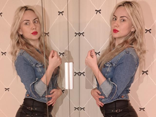 JenyShow has blonde hair, red lips, and wears a denim jacket with black pants. She's mirrored, showing her ass and hands on her hip. Her reflection is visible.