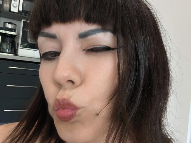 OrianaLaFrancaise with dark brown hair, black eyeliner, gray eyebrows, and pouty pink lips, making a kissing face, showing her soft, pale skin and slightly parted pink lips.