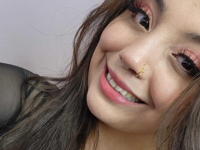 RoxannDiaz has a cute smile with pink eyeliner, long brown hair, and a gold nose ring, showing her teeth and wearing a black sheer top.