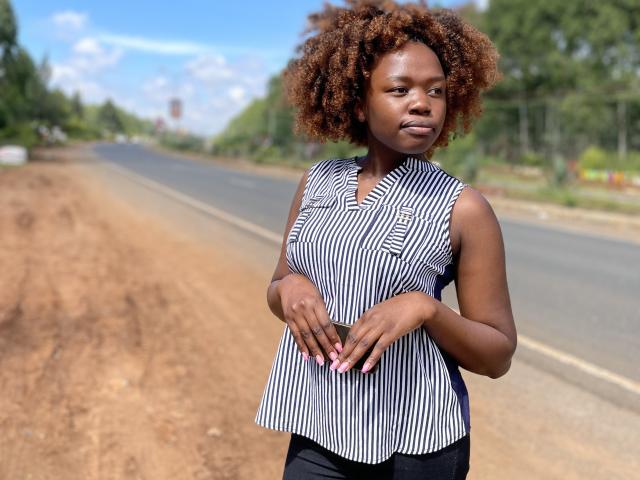 Yollah, dark-skinned with curly brown hair, wearing a sleeveless black and white striped top, stands on a dirt road, hands gently touching her belly, nails painted pink.