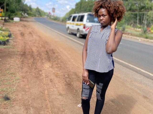 Yollah with dark brown skin, curly red-brown hair, wearing a striped sleeveless top and ripped black pants, stands on a dirt road, hand on head, looking serious.