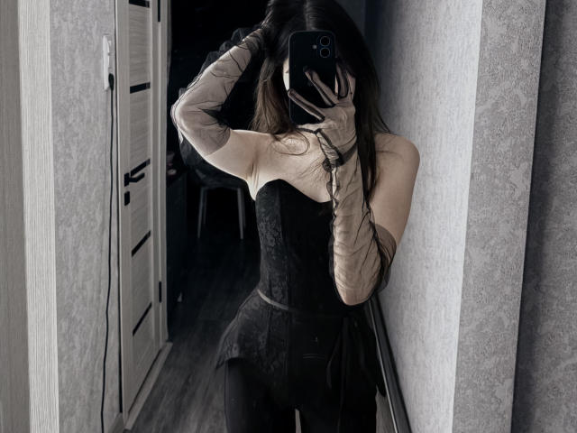 LezlieKennady with long brown hair, black corset, sheer gloves, and black pants, taking a mirror selfie, covering face with phone, in a dimly lit room.