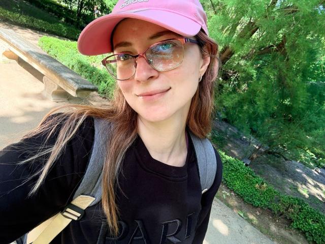 AmbeerWood has long brown hair, a pink cap, glasses, black shirt, and gray backpack, smiling with a cute nose and small ass, standing outdoors in a sunny, green park.