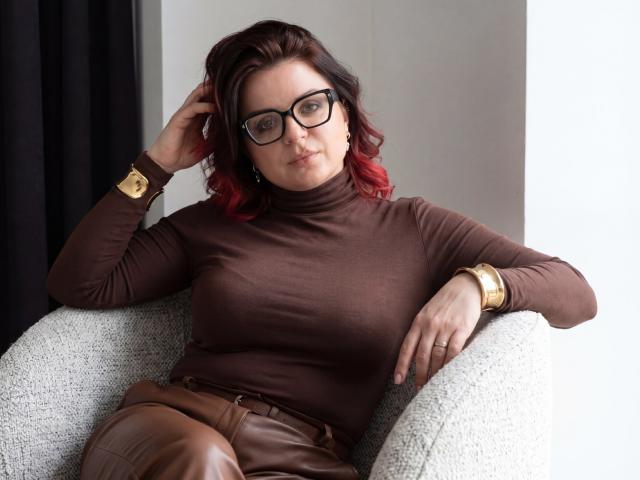 AmeliaDateri with red-tipped brown hair, black glasses, brown turtleneck, brown leather pants, gold bracelet, and gold watch, sitting in a white chair, hand in hair.