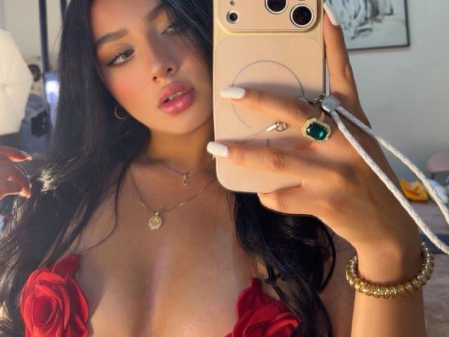 LillyTaylor with long black hair, red rose-patterned top showing her tits, holding a gold phone, wearing a green ring, white nails, gold bracelet, and a necklace.