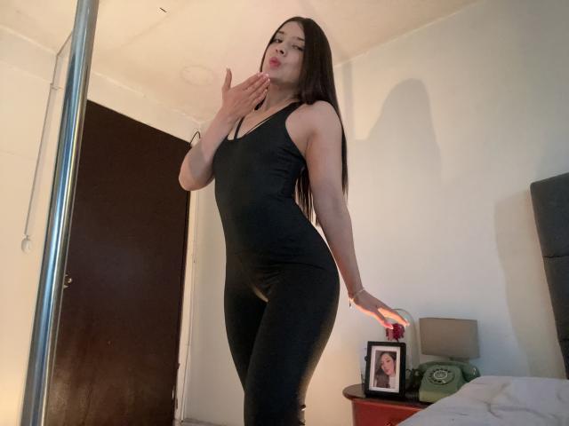 EmmaLombardi in a tight black dress, standing by a bed, hand on her lips, holding a photo frame, long dark hair, curvy, big ass, small tits, sexy, bedroom.