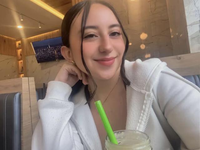 RosalieDupont with light brown hair, smiling, wearing a white zip-up hoodie, holding a drink with a green straw, in a wooden restaurant booth.