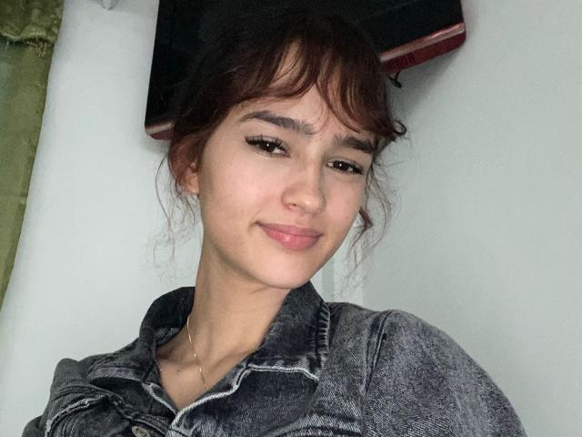 MelodyMansson has dark brown hair, a slight smile, and wears a denim shirt. She has a small nose, subtle makeup, and a cute, relaxed expression.