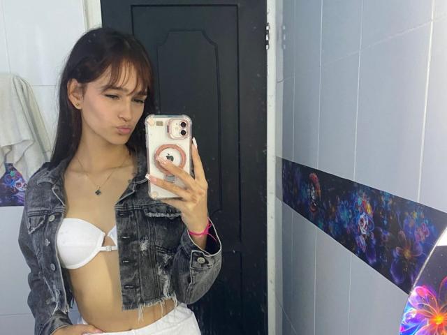MelodyMansson with long brown hair, wearing a white bra, denim jacket, and white pants, taking a selfie in a bathroom, showing her tits, holding an iPhone.