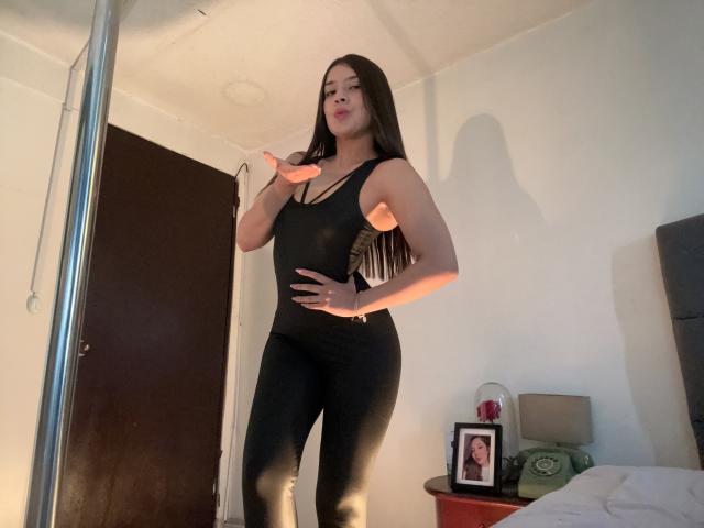 EmmaLombardi, standing in a room, wearing a tight black tank top and pants, showing off her toned arms and curvy ass, with long black hair and a confident smile.