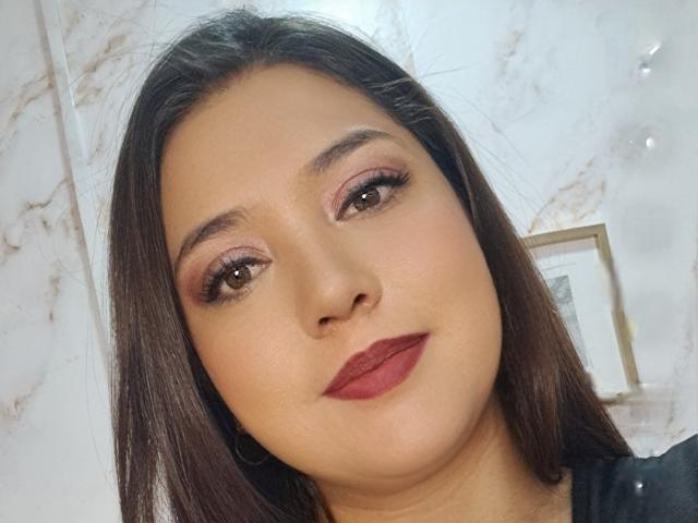 PaulaAnd has brown hair, pink eyeshadow, long lashes, and red lipstick. Her smooth tan skin and small nose are visible. She's wearing a black shirt.