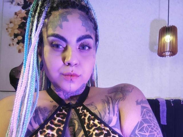 MilaBlake with colorful braids, tattoos, and nose ring, wearing leopard print top, has blood on her chin, and a pentagram tattoo on her arm, under purple lighting.