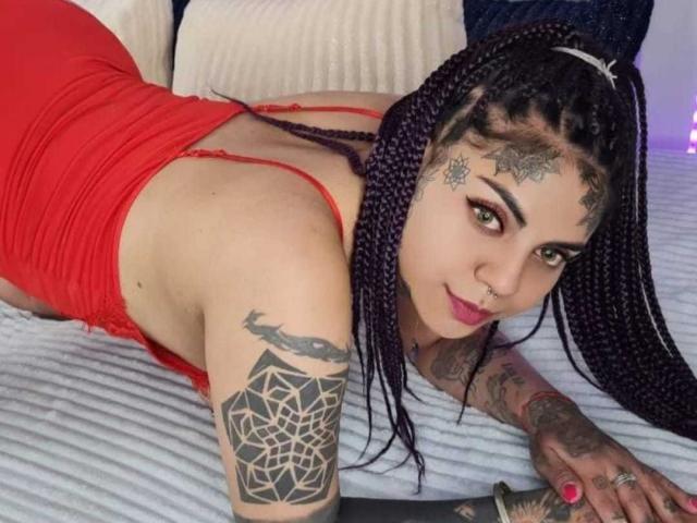 MilaBlake with purple braids, red dress, tattoos on arm and face, lying on bed, looking at camera, showing her ass, nose ring, and pink lipstick.