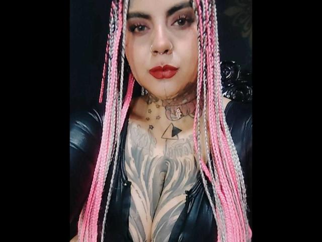 MilaBlake with pink braids, nose ring, red lipstick, black leather top, showing large chest tattoos, and a tattooed neck. Boobs, tattoos, pink braids, and nose ring are prominent.