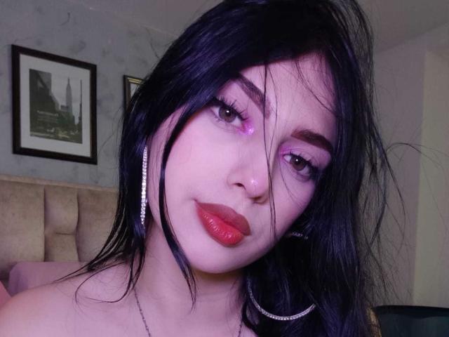 IsabellaaRossi has dark purple eyelids, red lipstick, black hair, and pinkish skin, wearing long silver earrings, with a subtle nose piercing and a slight smile.