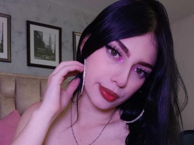 IsabellaaRossi with long black hair, purple eye makeup, red lipstick, silver hoop earrings, and a necklace, touching her ear, in a bedroom with framed black-and-white pictures.
