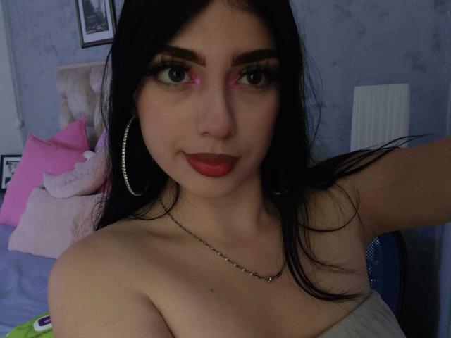 IsabellaaRossi has dark hair, green eyes with pink highlight, red lipstick, large hoop earrings, and a silver necklace. She's topless, showing her bare shoulders and chest.