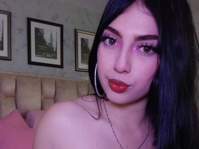 IsabellaaRossi with pink eyeshadow, red lipstick, black hair, hoop earrings, and bare shoulders, smiling at the camera, showing her tits slightly. Bedroom setting.
