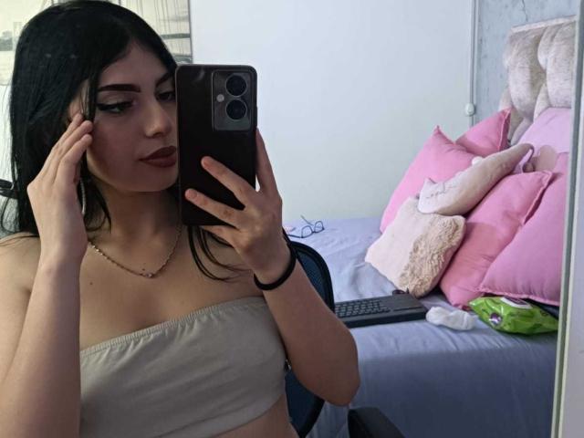 IsabellaaRossi with dark hair, wearing a strapless top, holding a phone, taking a mirror selfie, showing her tits, pink pillows, and a bed in the background.