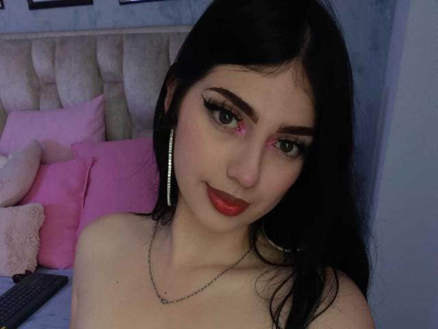 IsabellaaRossi with black hair, green eyes, red lipstick, pink eye makeup, silver hoop earrings, necklace, and red lipstick, looking at the camera, topless, in a dimly lit room.