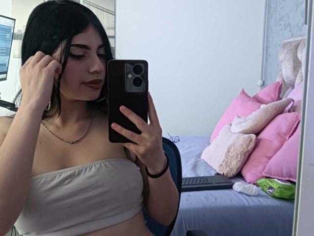 IsabellaaRossi with dark hair, wearing a strapless white top, holding a phone, taking a mirror selfie, showing her tits, pink pillows, and a white cat in the background.