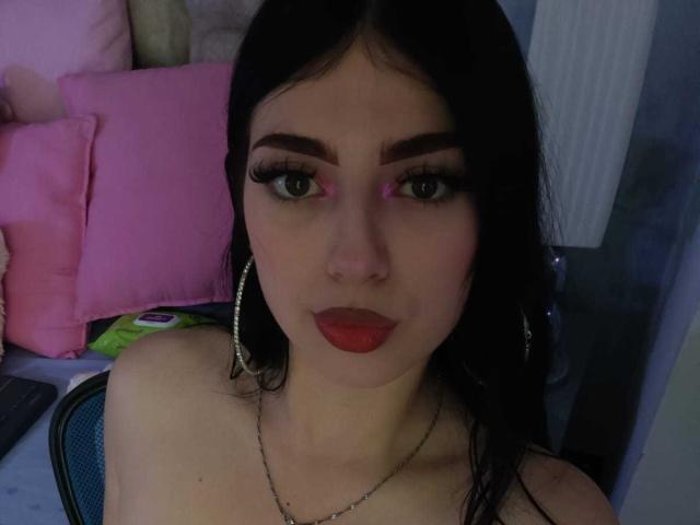 IsabellaaRossi with dark hair, green eyes, thick eyebrows, red lipstick, and large hoop earrings, wearing a necklace, sitting in a chair, looking at the camera.