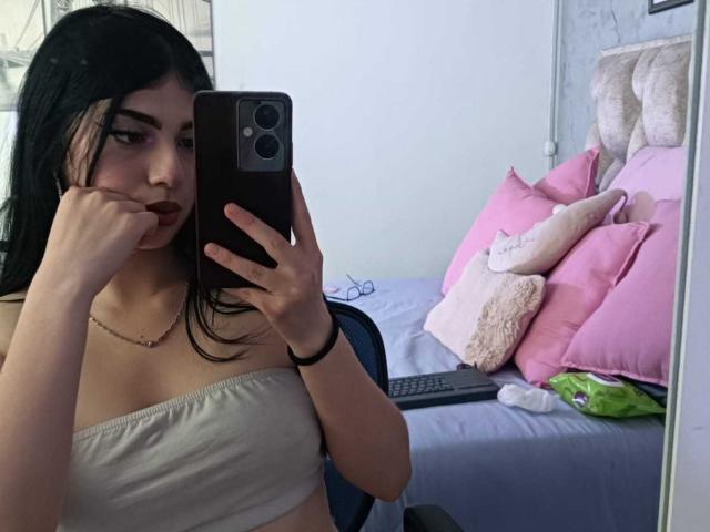 IsabellaaRossi with black hair, pouty lips, and a strapless white top, taking a mirror selfie with her phone, showing her small tits and a necklace.