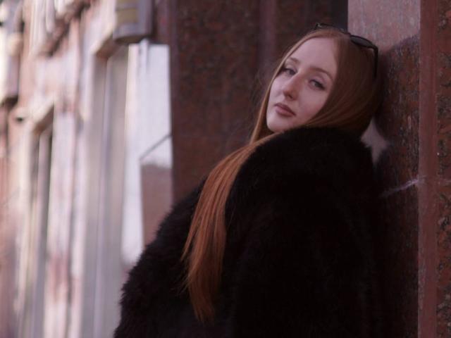DeloraBangert with long red hair, wearing a black fur coat, leaning against a brick wall, looking over her shoulder with a slight smile. Eyeglasses rest on her head.