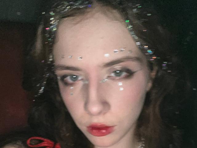 EviaMarcot has glitter on her forehead and cheeks, with sparkly hair, red lips, and thick black eyeliner. Her blue eyes are slightly squinted. She's looking at the camera.