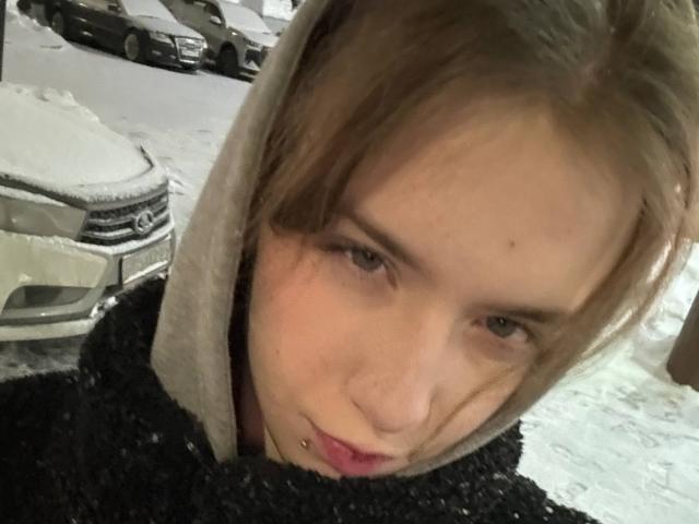 EviaMarcot has a pouty face, light brown hair, and a nose piercing, wearing a black textured hoodie with a beige hood, snow on a car in the background.
