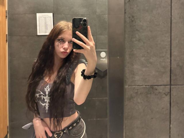EviaMarcot with long brown hair, gray crop top, black scrunchie, and black belt, holding phone in mirror, showing small tits and slim waist, standing in bathroom.