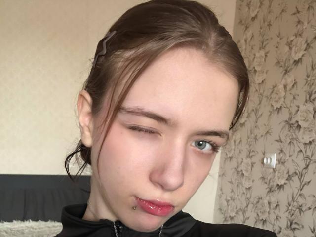 EviaMarcot has pale skin, brown hair in a braid, winks one blue eye, has pink lips with a bottom lip piercing, and wears a black zip-up top.