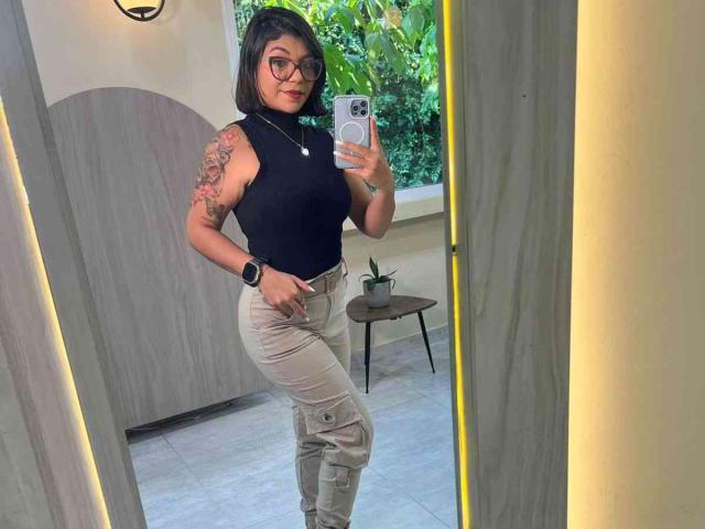 Valery Pierce, com cabelo preto curto, óculos, blusa preta sem mangas, calças bege, braço tatuado, segurando um celular, usando um relógio, em pé em frente a um espelho, com uma planta verde ao fundo..