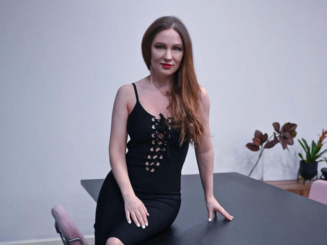 DashaHartt-hot with long brown hair, black lace-up dress, sitting on black table, smiling, red lipstick, pale skin, white nails, pink chair, potted plant in background.