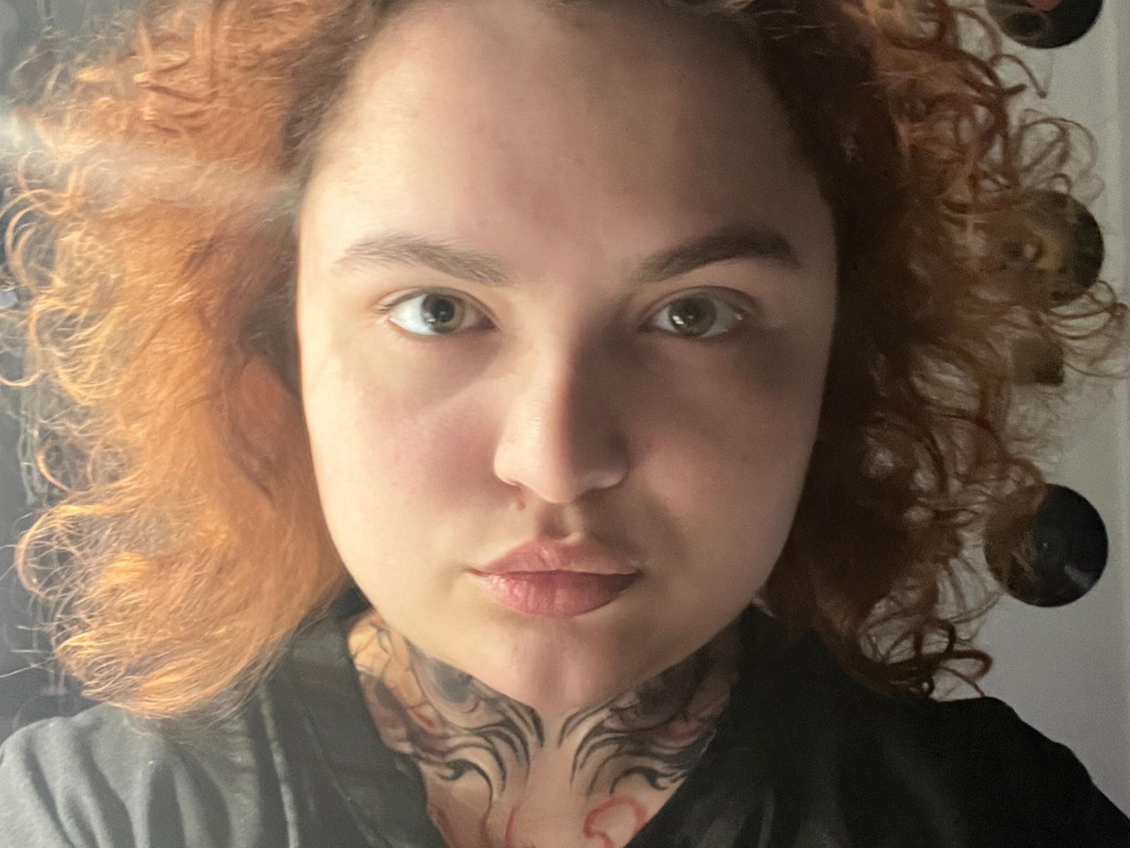 AnjelinaSweet with curly red hair, tattoos on her neck, green eyes, and a serious expression, wearing a gray shirt, looking directly at the camera.