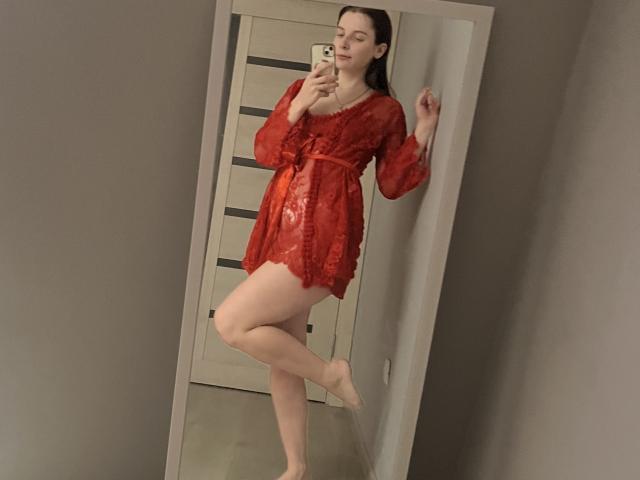 Meliss69 trägt ein rotes Spitzenkleid, steht auf einem Bein, hält ein Telefon vor einem Spiegel und hat nackte Füße und langes, dunkles Haar. Ihre Brüste sind sichtbar..