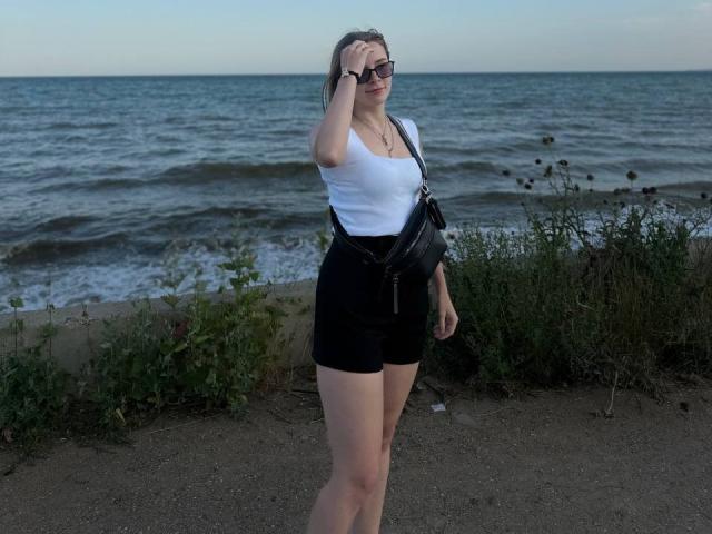 Meliss69 heeft bleke huid, draagt een witte tanktop, zwarte shorts en een zwarte schoudertas, staat bij de oceaan, met haar hand op haar voorhoofd, en glimlacht..