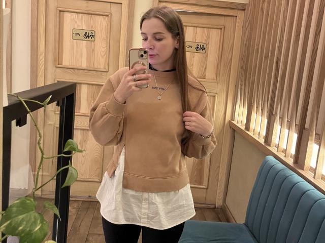 Meliss69 with long brown hair, wearing a tan sweater, white skirt, black choker, and black pants, taking a mirror selfie in a wooden room, holding a phone.