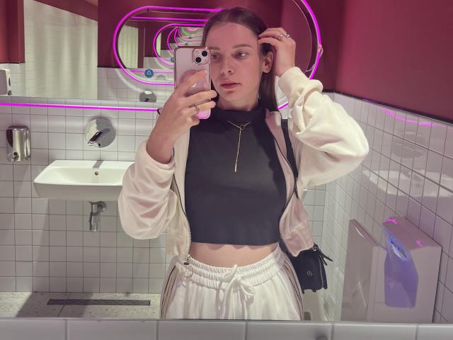 Meliss69, avec une peau claire, des cheveux bruns, portant un crop top noir, une veste blanche et un pantalon blanc, tenant un téléphone, éclairée par des lumières roses néon, dans une salle de bain, montrant sa poitrine plate..