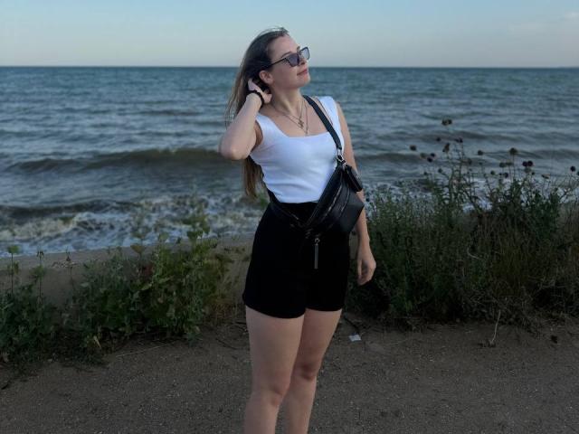 Meliss69 mit langen, braunen Haaren, die schwarze Shorts und ein weißes Tanktop trägt, eine schwarze Umhängetasche und eine Sonnenbrille, steht am Strand, eine Hand im Haar, und blickt nach oben..