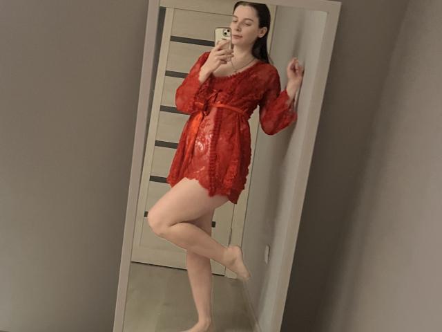Meliss69, vêtue d'une robe rouge en dentelle, debout sur une jambe, prend un selfie devant un miroir avec son téléphone, montrant ses fesses et ses seins nus, avec de longs cheveux bruns et une peau pâle..