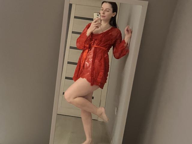 Meliss69, com as pernas descobertas, vestindo um vestido vermelho de renda e com cabelos longos e escuros, está em pé sobre uma perna, tirando uma selfie no espelho com um celular, mostrando seus seios e o vestido vermelho..