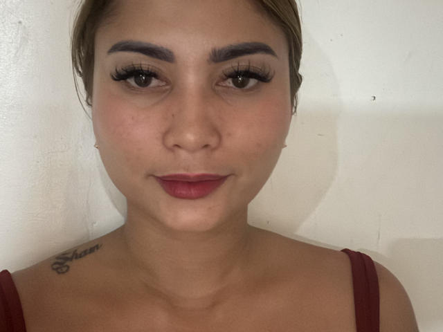 KareyGrasman has tan skin, brown eyes, thick eyebrows, long eyelashes, red lipstick, and a small "Stain" tattoo on her right shoulder. She's wearing a red top.