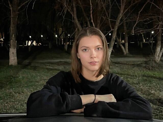 JennellMatacale with straight brown hair, green eyes, and a serious expression, wearing a black sweater, arms crossed, standing in a dark, wooded area at night.