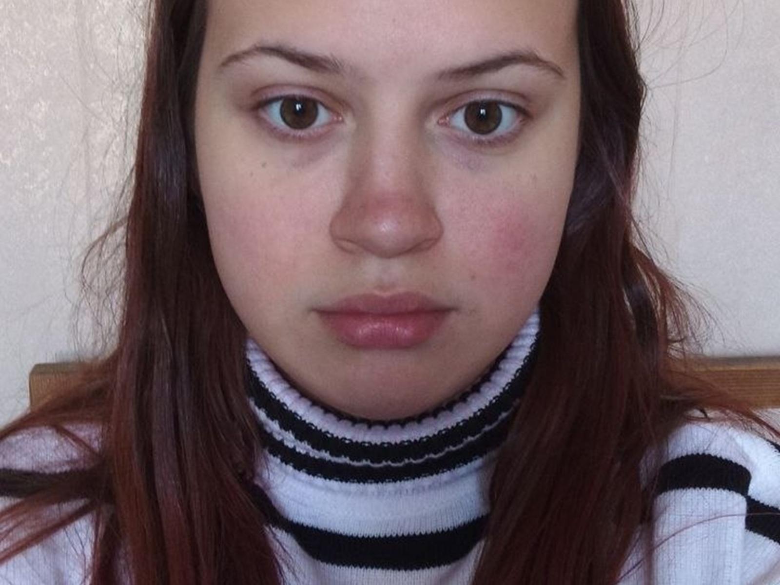 TaishaGloden ha la pelle chiara, gli occhi marroni e i capelli rossi, indossa un maglione a collo alto bianco con strisce nere, appare seria con le guance leggermente rosate e le labbra leggermente serrate..