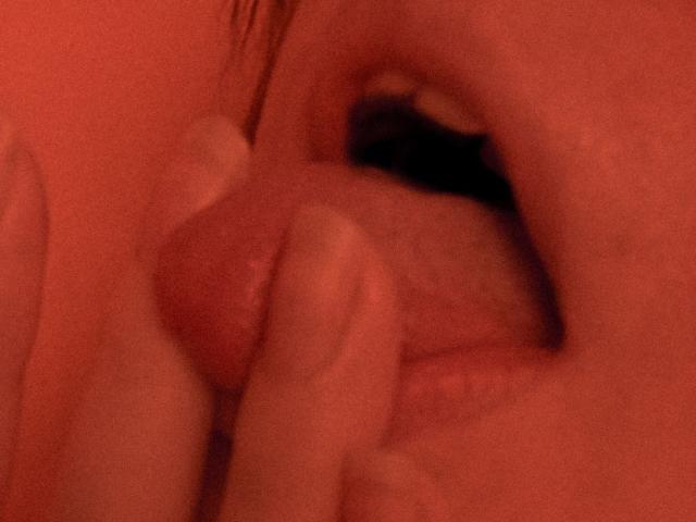 AvaNikol's mouth is open, with her tongue out, and a finger gently holding her pink, erect, circumcised penis. The image is red-lit.