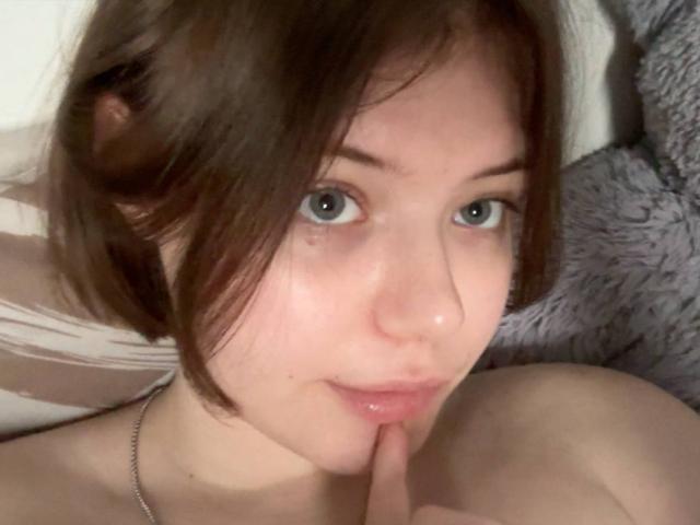 AvaNikol with short brown hair, blue eyes, and fair skin, biting her finger, wearing a necklace, looking directly at the camera, showing her bare shoulders.