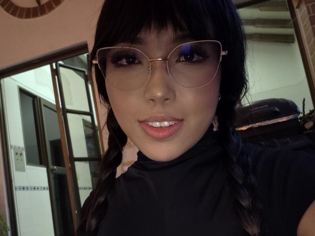 Heyly with black hair in braids, wearing glasses, and a black turtleneck, showing a subtle smile with shiny pink lips. Her eyes are dark, and she has a cute face.